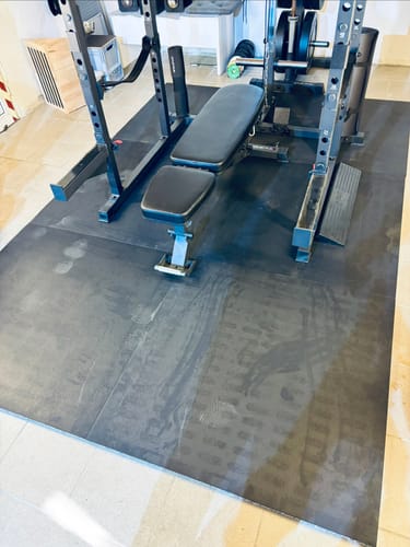 Ein Kunde zeigt die verlegten MaxProtect Bodenschutzmatten, auf denen eine Hantelbank und ein Power-Rack stehen.