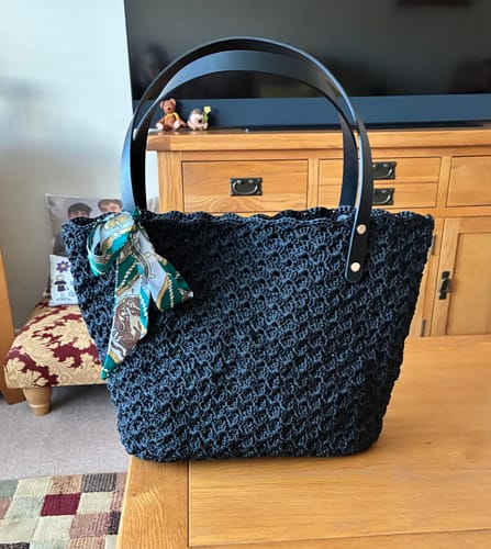 Customer's completed black Aveline crochet Shoulder Bag with a decorative scarf tied to the leather strap, sitting on a table.