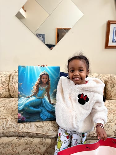 A happy child smiling next to her personalized The Mermaid Princess canvas portrait on a couch.
