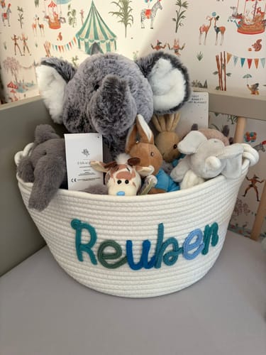 Reviewer's white personalized knitted basket with 'Reuben' in multicolor yarn, filled with stuffed animals in a crib.