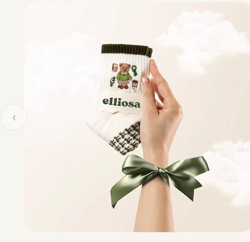 A reviewer's hand holding up a white Merry Beary sock with a cute bear design and green trim.