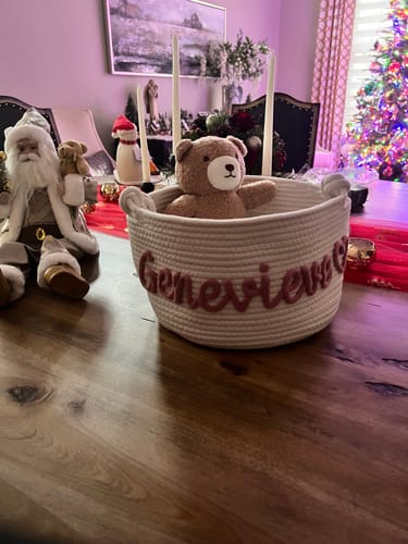 Customer's personalized white knitted basket with the name 'Genevieve' in pink yarn, holding a teddy bear on a table.