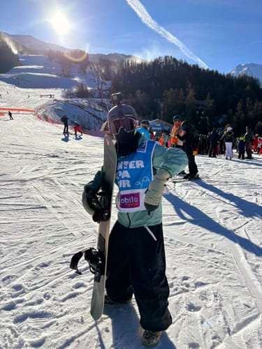 Reviewer on a ski slope wearing the baggy black snow pants while holding a snowboard.
