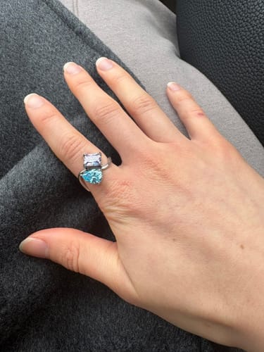 A customer's hand wearing the silver Navae Birthstone Ring, which features a light blue pear-cut stone and a pale purple radiant-cut stone.