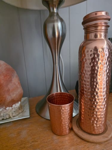 Customer's hand-hammered Pure Copper Water Bottle and matching cup sitting on a wooden surface next to a lamp.