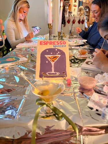 Reviewers at a fun sip and paint party, with a finished 'Espresso Martini' mini painting from the kit displayed on the table.