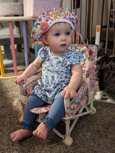 Customer's baby wearing a matching floral hat while sitting in the Paloma portable booster chair indoors.