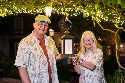 Two reviewers smiling in matching Mickey's Christmas Carol shirts at night while holding a decorative lantern.