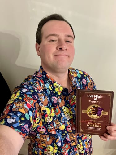 Customer wearing the colorful Five Nights at Freddy's 'Fantasy and Fun' shirt while holding a book.