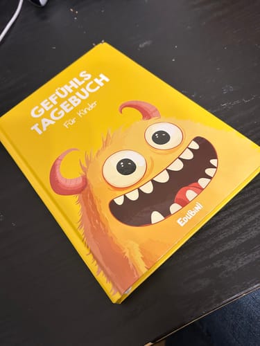 Ein Rezensent zeigt das gelbe Gefühlstagebuch für Kinder mit einem lustigen Monster-Cover, das auf einem Tisch liegt.