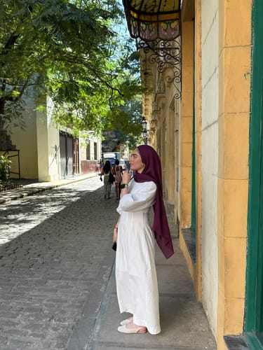 Customer standing on a cobblestone street wearing a long white dress and a lovely dark red hijab.