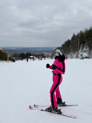Reviewer on a ski slope wearing the slim-fit fuchsia one-piece ski suit with a black faux fur hood.