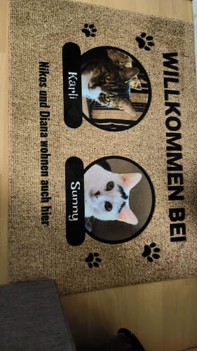 Kunde zeigt personalisierte Fußmatte mit Fotos der Katzen Karli und Sunny, die auf einem Holzboden liegt.