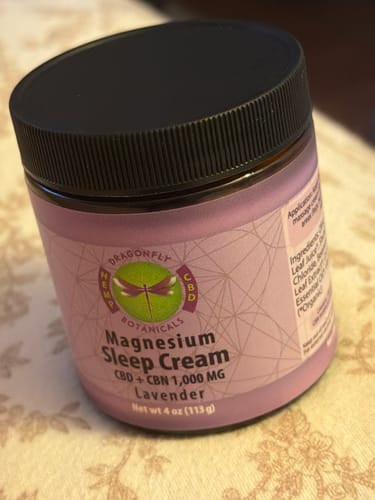 Customer's jar of CBD + CBN Magnesium Sleep Cream in Lavender, resting on a floral-patterned surface.