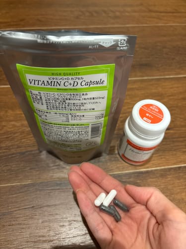 レビューアーの手のひらに乗った白い「ビタミンC+Dカプセル」と他のカプセル。背景に商品のパッケージ。