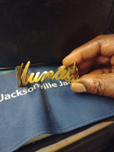 A customer's hand holding a gold Personalized Name Bangle with the name 'Numac' crafted into the band.