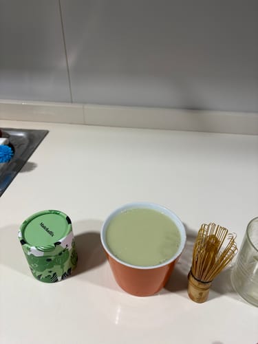 Cliente muestra su té matcha preparado en una taza naranja, junto al envase del producto y un batidor de bambú.