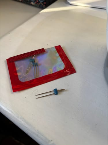Customer's Singer twin needle on a white surface in front of its iridescent red packaging.