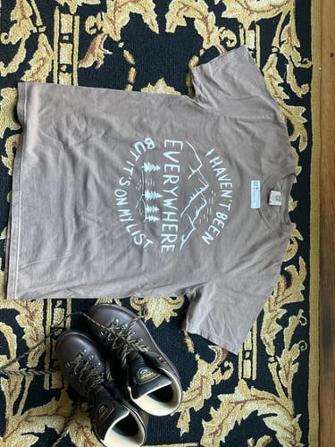 Customer's brown "Haven't Been Everywhere T-shirt" laid flat on a rug next to a pair of hiking boots.