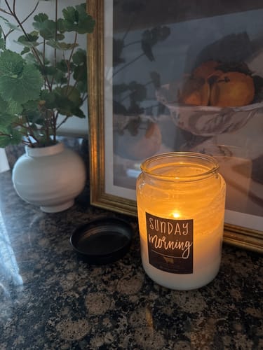 Customer's lit Sunday Morning Candle in a glass jar, providing a warm glow on a dark countertop.