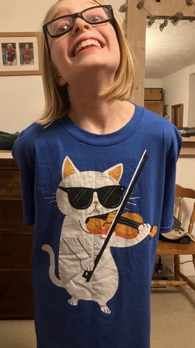 A young reviewer smiles while wearing the blue Cat Playing Violin T-Shirt, which has a slightly oversized fit.
