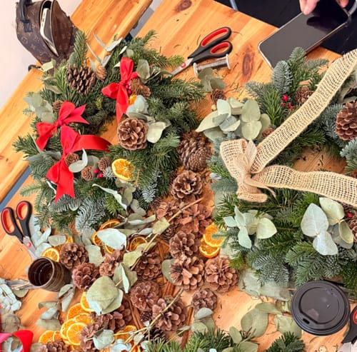 Customer's finished wreaths from the Wreath Making Workshop, surrounded by pine cones, dried oranges, and crafting materials.