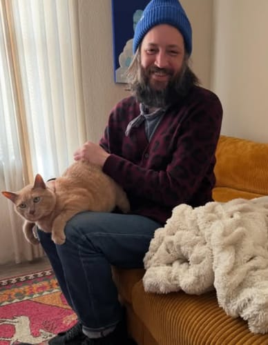 Customer smiling while sitting on a couch with a cat on their lap, wearing a blue hat.