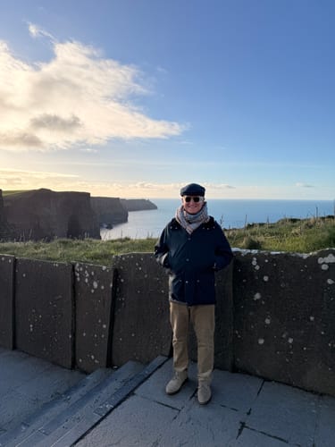 Reviewer wearing the dark Vintage Cap Tweed while standing outdoors with coastal cliffs in the background.