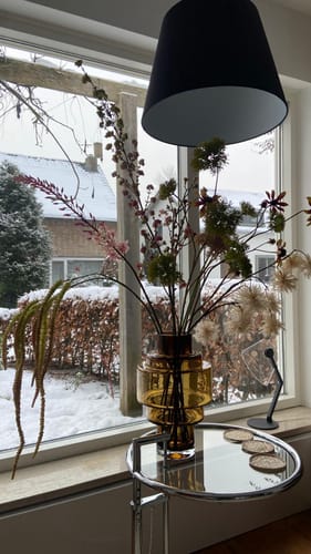 Recensent toont de cognackleurige Kasai M vaas gevuld met een boeket bloemen op een bijzettafel voor een raam.