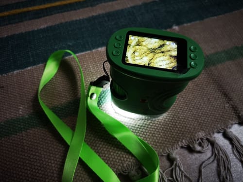 Rezensent zeigt das grüne Edubini Abenteuer-Mikroskop mit eingeschaltetem Bildschirm und Licht auf einer Decke.