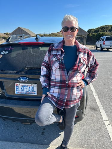Reviewer smiling outdoors wearing the blue Ainsley Sweater layered under a plaid jacket near the beach.