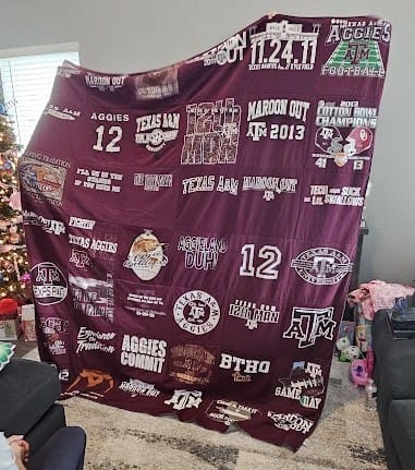 Customer holding up their large, maroon Premium T Shirt Quilt made from various Texas A&M University shirts in a living room.