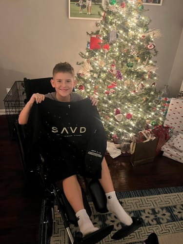 A smiling customer in a wheelchair holds their black Youth Oversized Hoodie in front of a Christmas tree.