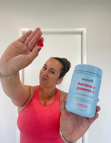 Reviewer holding a blue container of Moodi hormone gummies and a single red, heart-shaped gummy.