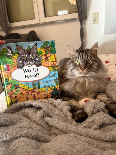 Kunde zeigt das personalisierte Wimmelbuch neben seiner Katze, die der Illustration auf dem Cover sehr ähnlich sieht.