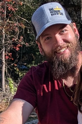 A customer smiling while wearing the gray and black Dad, Husband, Man of God trucker hat outdoors.
