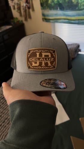 Customer holding a grey and black trucker-style custom hat with an engraved leather patch showing the initials 'JR' and 'DELALUZ'.