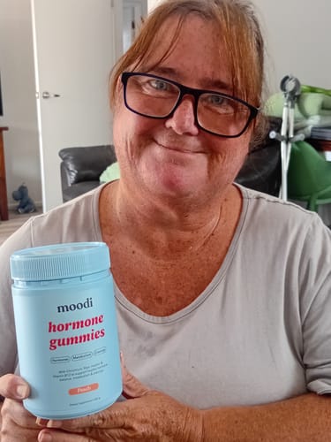 Reviewer smiling while holding a light blue jar of Peach hormone gummies in their home.