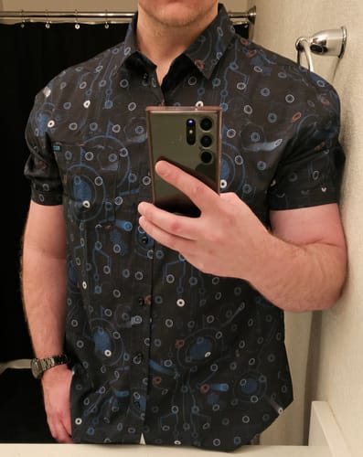 Reviewer taking a mirror selfie to show the fit of the black, patterned Tron Legacy KUNUFLEX shirt.