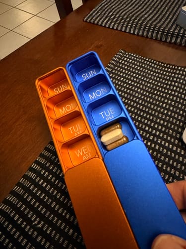 Customer holding two metal Weekly AM-PM Vitamin Cases, one orange and one blue, open to show the daily compartments.