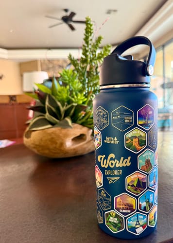 Customer's dark blue Lets Wander World Explorer Water Bottle on a table, showing the colorful destination stickers applied.
