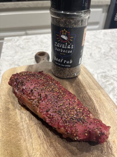 Customer's raw steak on a wooden board, heavily seasoned with Zavala's Beef Rub next to the shaker bottle.
