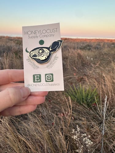 Reviewer holding the custom rabbit skull pin in a field with the sunset in the background.