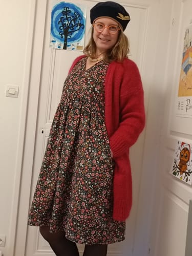 Une cliente portant la robe Edonie à fleurs, avec un gilet rouge et un béret, la main dans la poche.