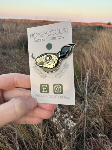 Customer holding a detailed enamel pin of a rabbit skull on its backing card outdoors.