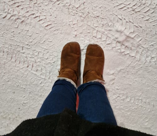 Arvostelijan ruskeat Vaistoa Huurre -paljasjalkabootsit jalassa lumisella alustalla.