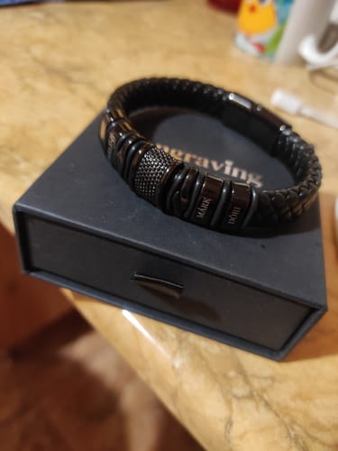 Customer's personalized black braided bracelet with engraved name charms resting on its product box.
