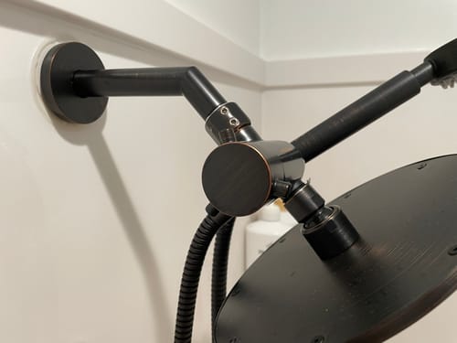 Close-up of a reviewer's dark 7 Inch Shower Arm and Flange mounted on a white shower wall.