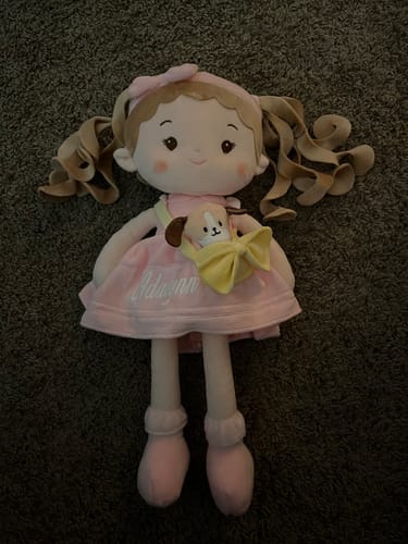 Customer's soft, personalized Milly Girl doll in a pink dress with her pet dog in a yellow pouch, lying on a dark carpet.
