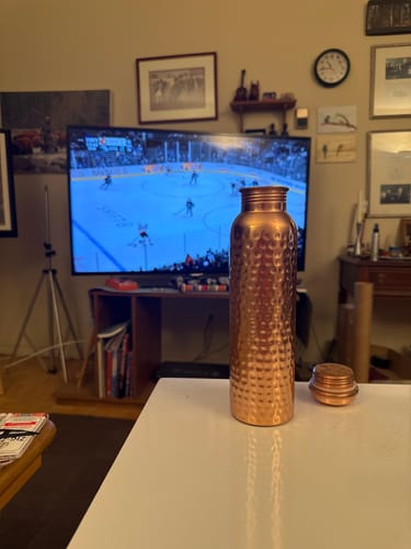 Customer's hand-hammered Pure Copper Water Bottle on a white table in a living room, with its cap beside it.